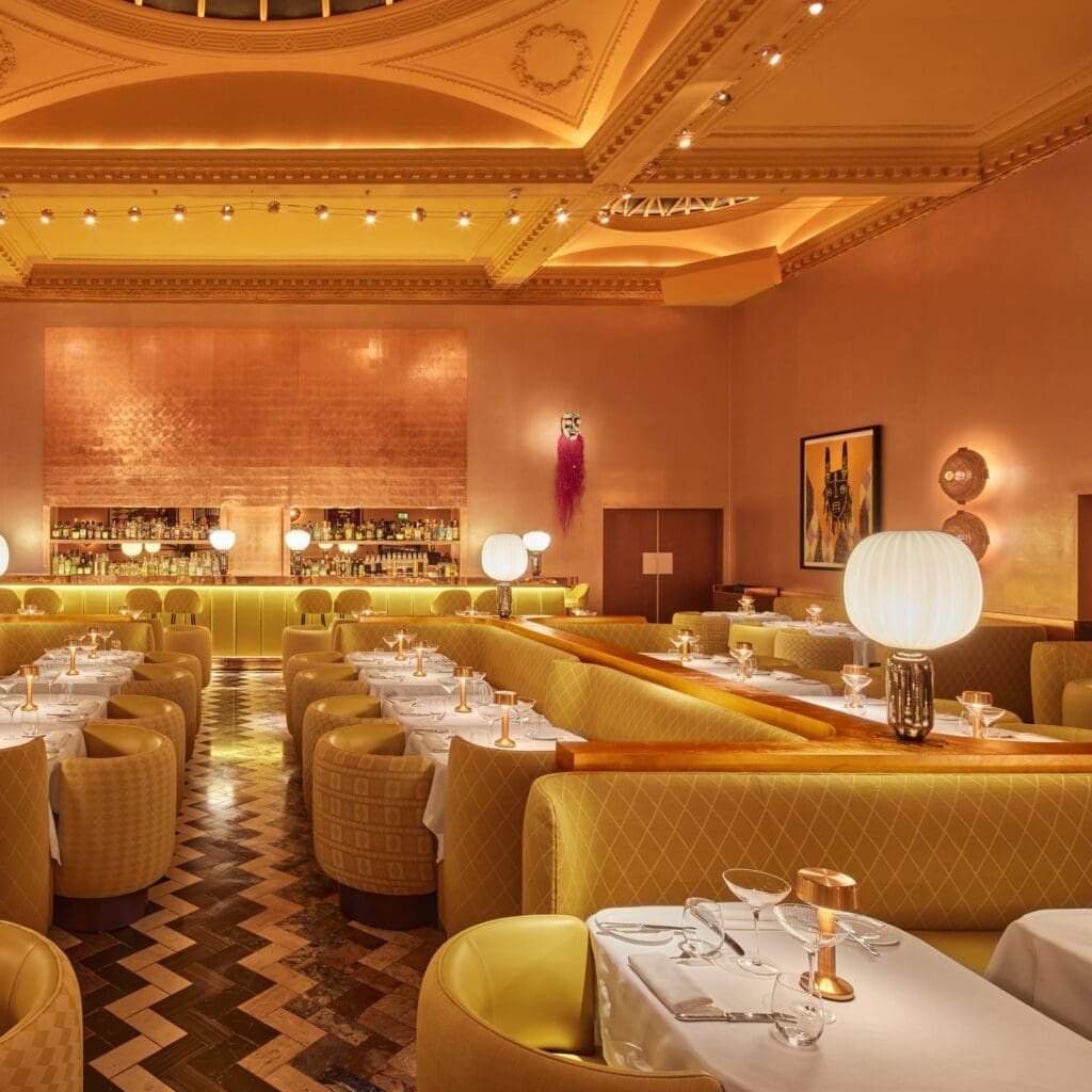 Off-axis view of the Gallery at Sketch from beside a set table, yellow tub chairs and banquettes leading to the lit bar under the copper-tiled wall, a white globe lamp and the ornate cornice above