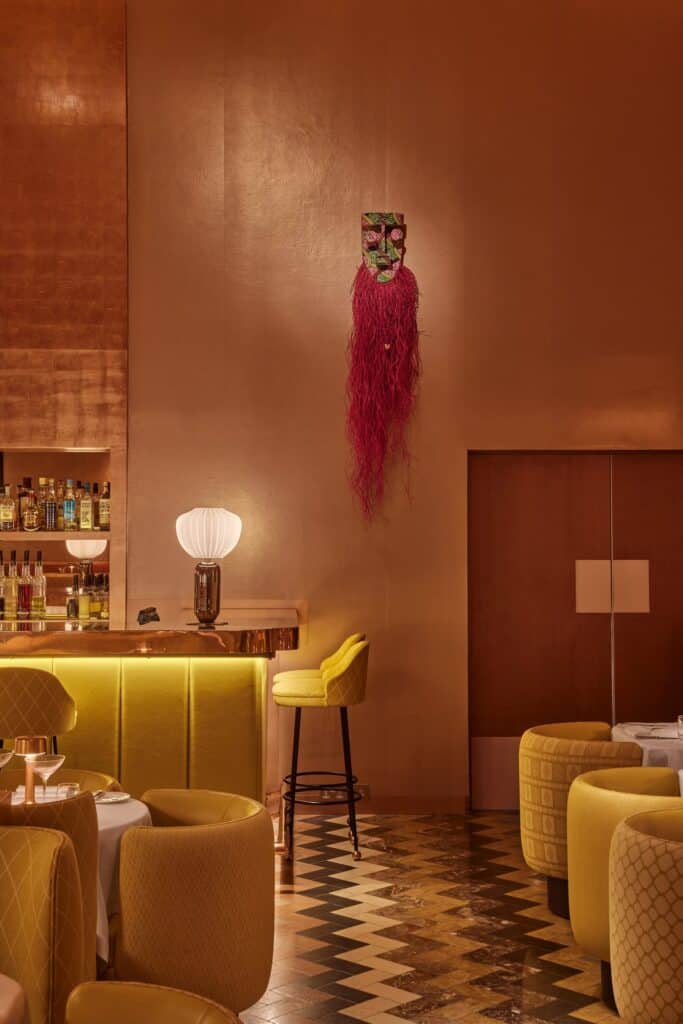 Corner of the Gallery at Sketch by the bar, a carved mask with a long pink raffia fringe on the copper wall above a yellow counter, tub chairs on a chevron-patterned floor beside a timber door