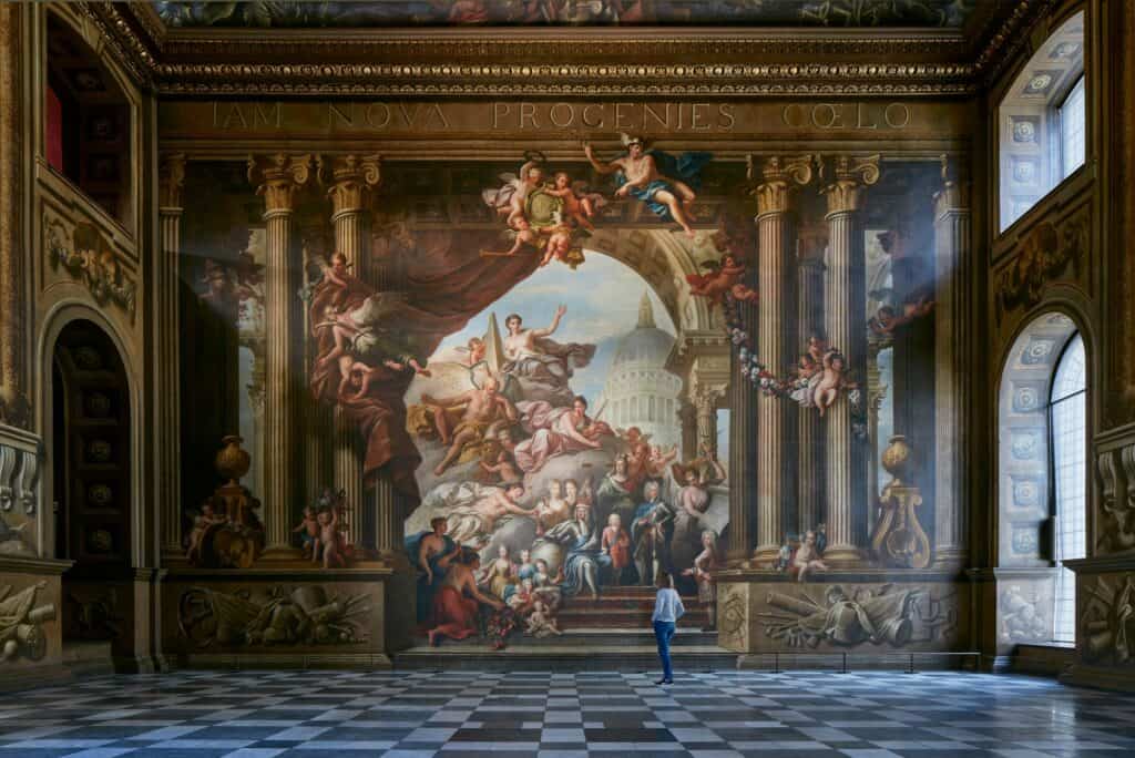 Thornhill's painted west wall in the Upper Hall of the Painted Hall, a crowd of allegorical figures beneath a painted arch and dome framed by fluted pilasters, with a lone visitor on the black and white marble floor