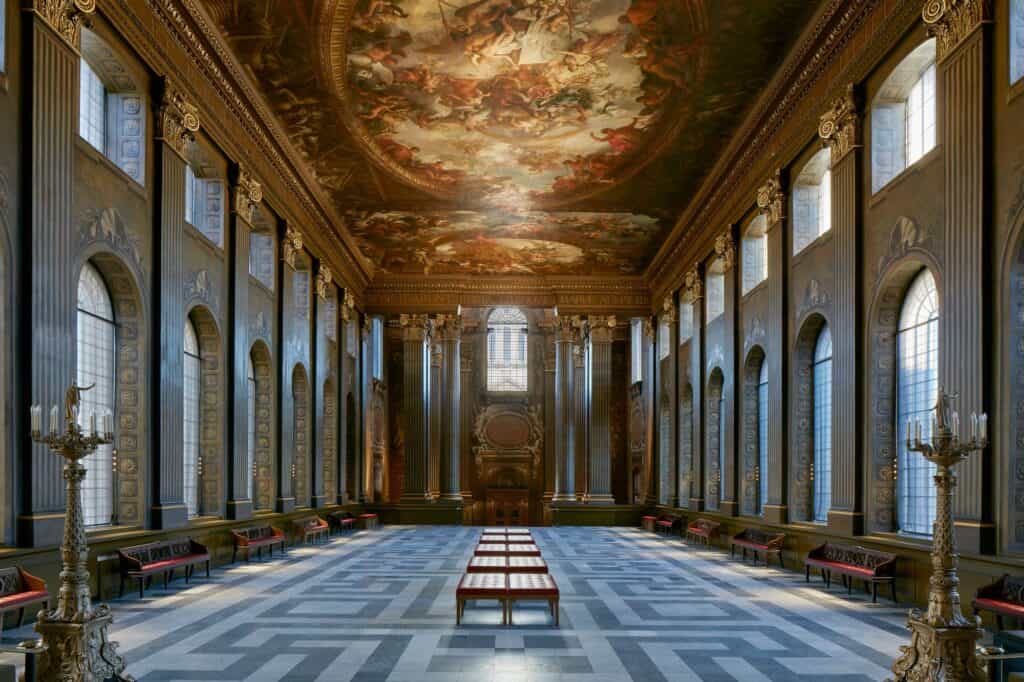 Symmetrical view down the length of the Painted Hall in Greenwich, gilded candelabra in the foreground, red-cushioned benches on a grey and white geometric marble floor and the painted oval ceiling overhead