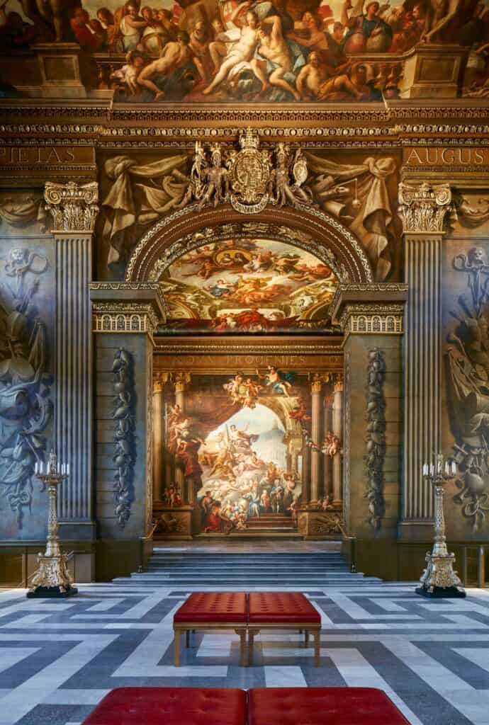 Gilded arch into the Upper Hall of the Painted Hall, a royal coat of arms crowning painted columns and the west wall mural, with red leather benches and tall candelabra in front