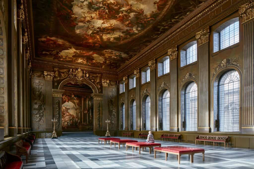 Angled view along the Painted Hall with sunlight falling through the arched windows onto red benches, a visitor seated beneath the restored ceiling and the Upper Hall arch beyond