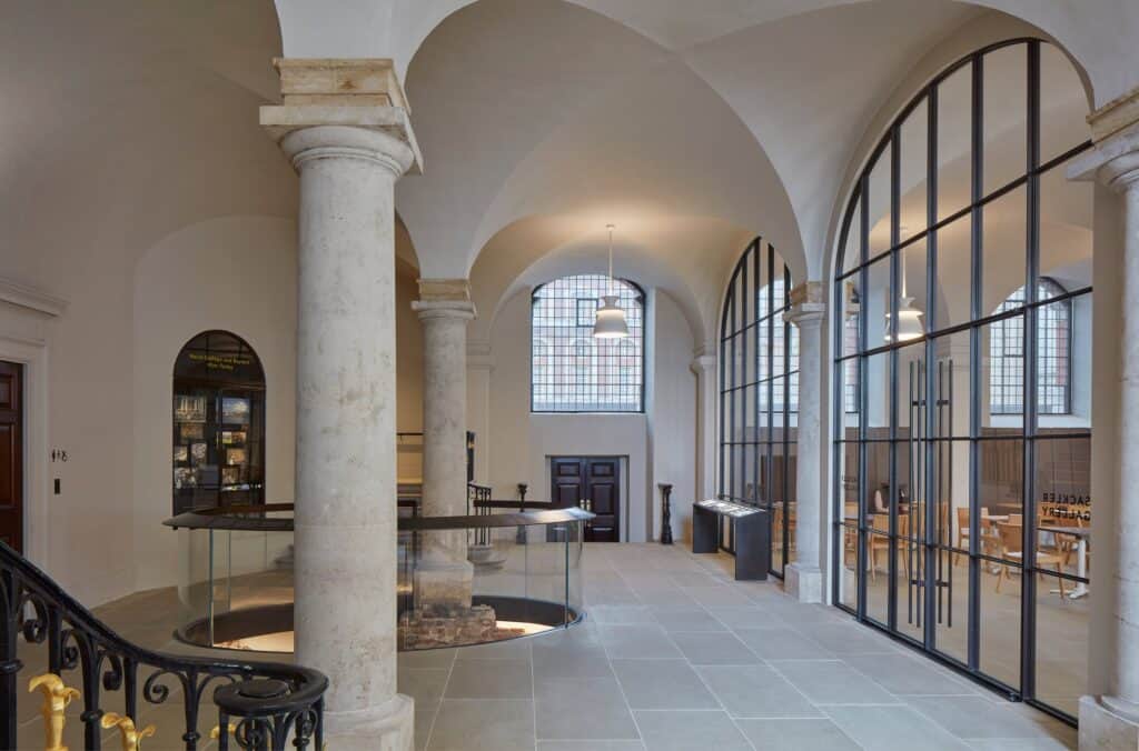 King William Undercroft below the Painted Hall with a stone column, a black and gilt stair balustrade, an oval glass opening in the stone floor and a tall bronze-framed glazed screen under white vaults