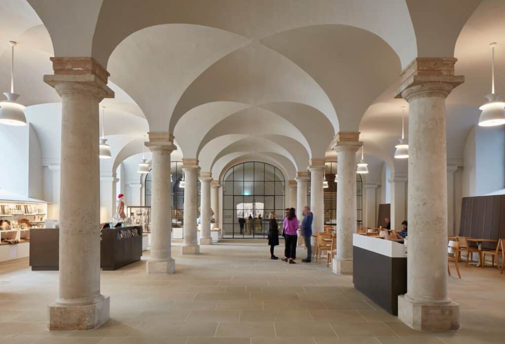 Painted Hall café in the King William Undercroft, a colonnade of stone columns carrying white groin vaults over a pale stone floor, with a dark serving counter and pendant lights