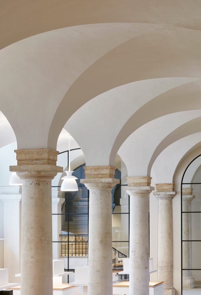 Stone columns with weathered capitals receding beneath curving white vaults in the Painted Hall undercroft, a white pendant lamp and a glazed screen with a stair beyond