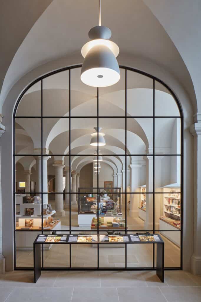 Arched bronze-framed glazed screen in the undercroft of the Painted Hall, a white pendant lamp in front and the shop's display shelves and vaulted aisle visible through the glass