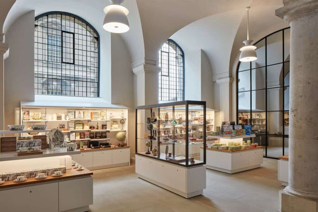Painted Hall shop in the undercroft, white display counters and a glass cabinet of globes and gifts beneath the vaults, with tall arched gridded windows and a stone column
