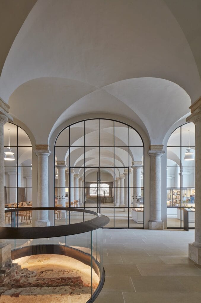 Oval opening edged with a curved glass and bronze balustrade revealing brick remains of the Tudor Greenwich Palace beneath the undercroft vaults, a glazed arch to the Sackler Gallery behind