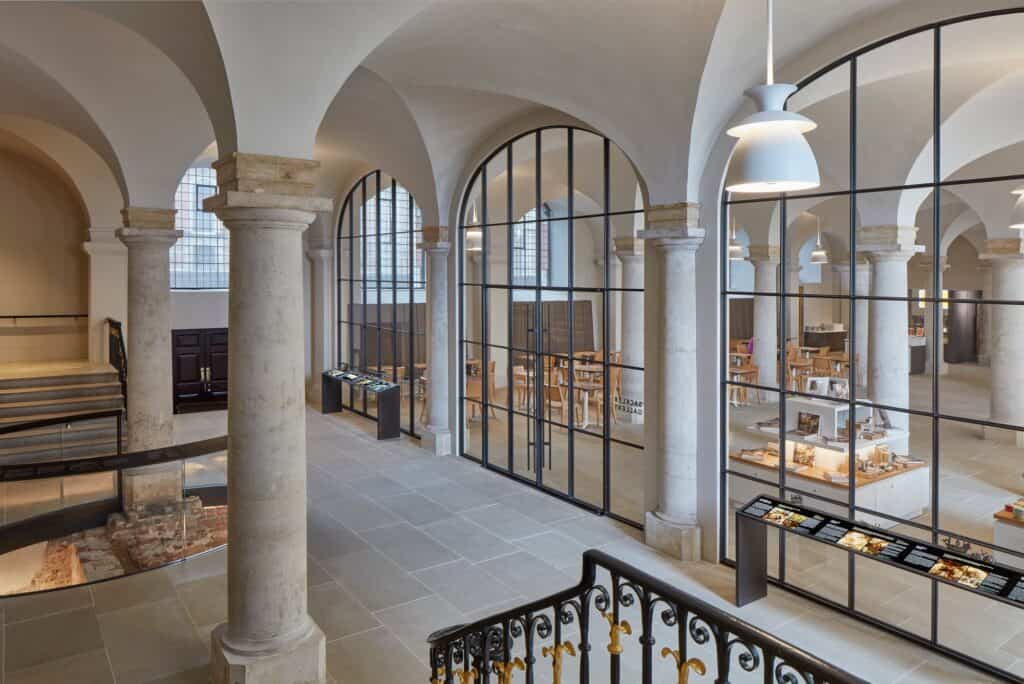 View from the gilded stair balustrade into the Painted Hall undercroft, stone columns and bronze-framed glazed screens dividing the café from the foyer, with the palace remains glimpsed at left