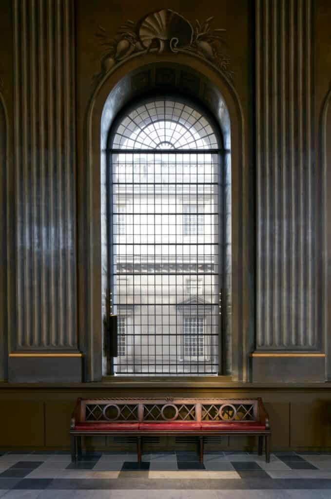 Tall arched window with a carved shell above, framed by fluted grey pilasters in the Painted Hall, a red upholstered bench with a circle-patterned back on the marble floor below