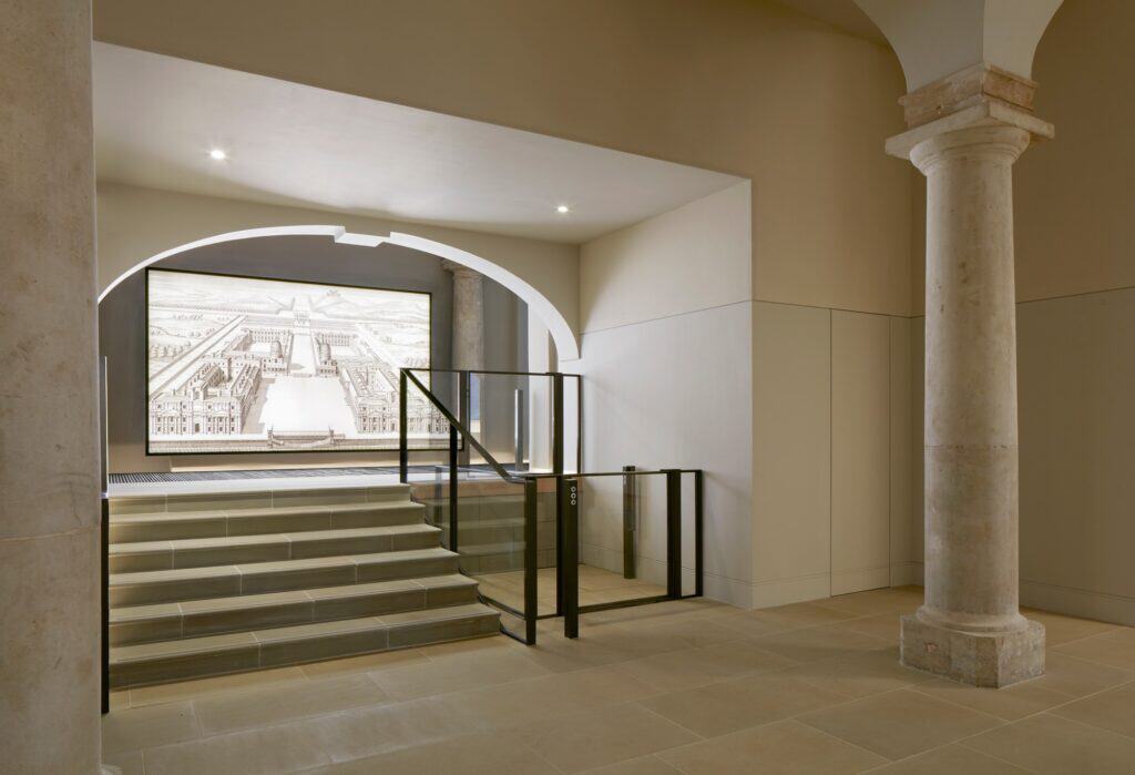 Stone steps with black steel railings rising through an arch in the Painted Hall undercroft towards a screen showing a historic bird's-eye engraving of Greenwich, beside a stone column