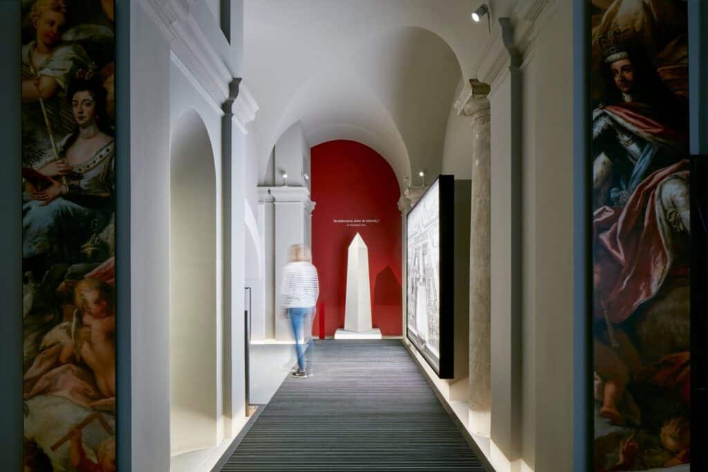 Interpretation corridor in the Painted Hall undercroft lined with large prints of Thornhill's painted figures, a dark carpeted floor leading to a white obelisk against a red wall, with a visitor walking through