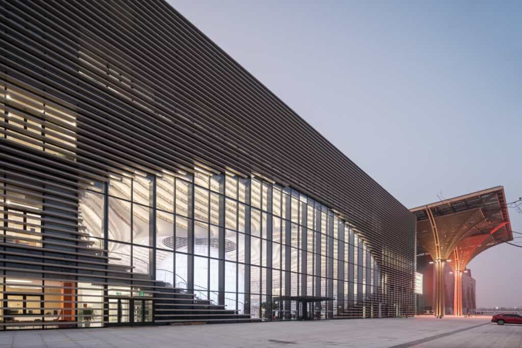 Corner of the louvred Tianjin Binhai Library facade at dusk beside a vaulted canopy of the cultural district