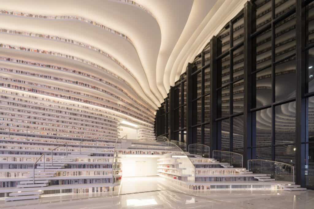 Lit bookshelf terraces and glass balustrades beside tall black-framed windows at Tianjin Binhai Library in the evening